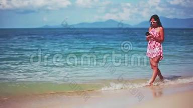 美丽的黑发女郎穿着红色连衣裙和太阳镜在沙滩上散步，使用手机。 <strong>1920</strong>x1080